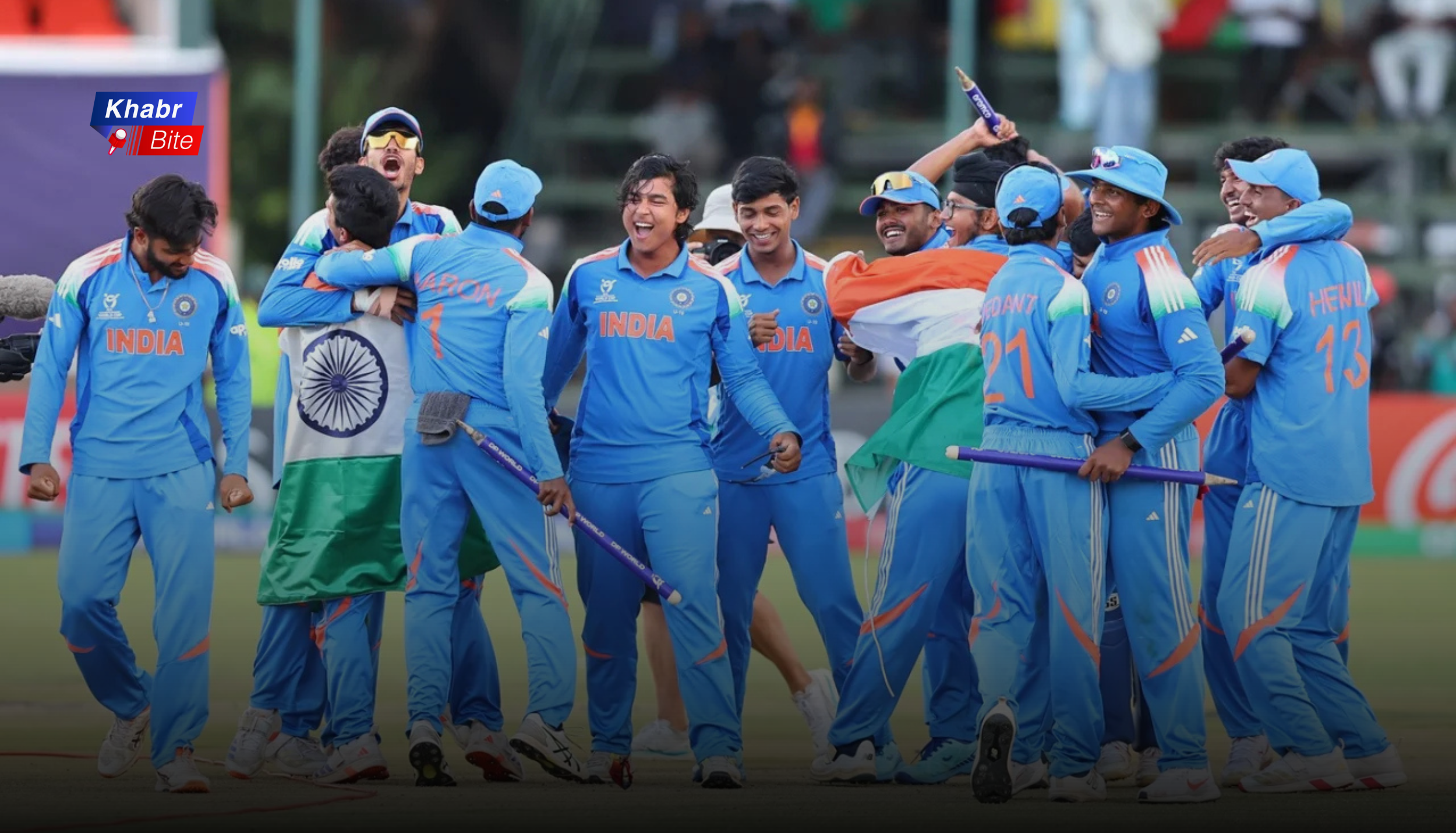India Under-19 cricket team celebrating a victory on the field, players in blue jerseys cheering and hugging while waving the Indian tricolour flag.