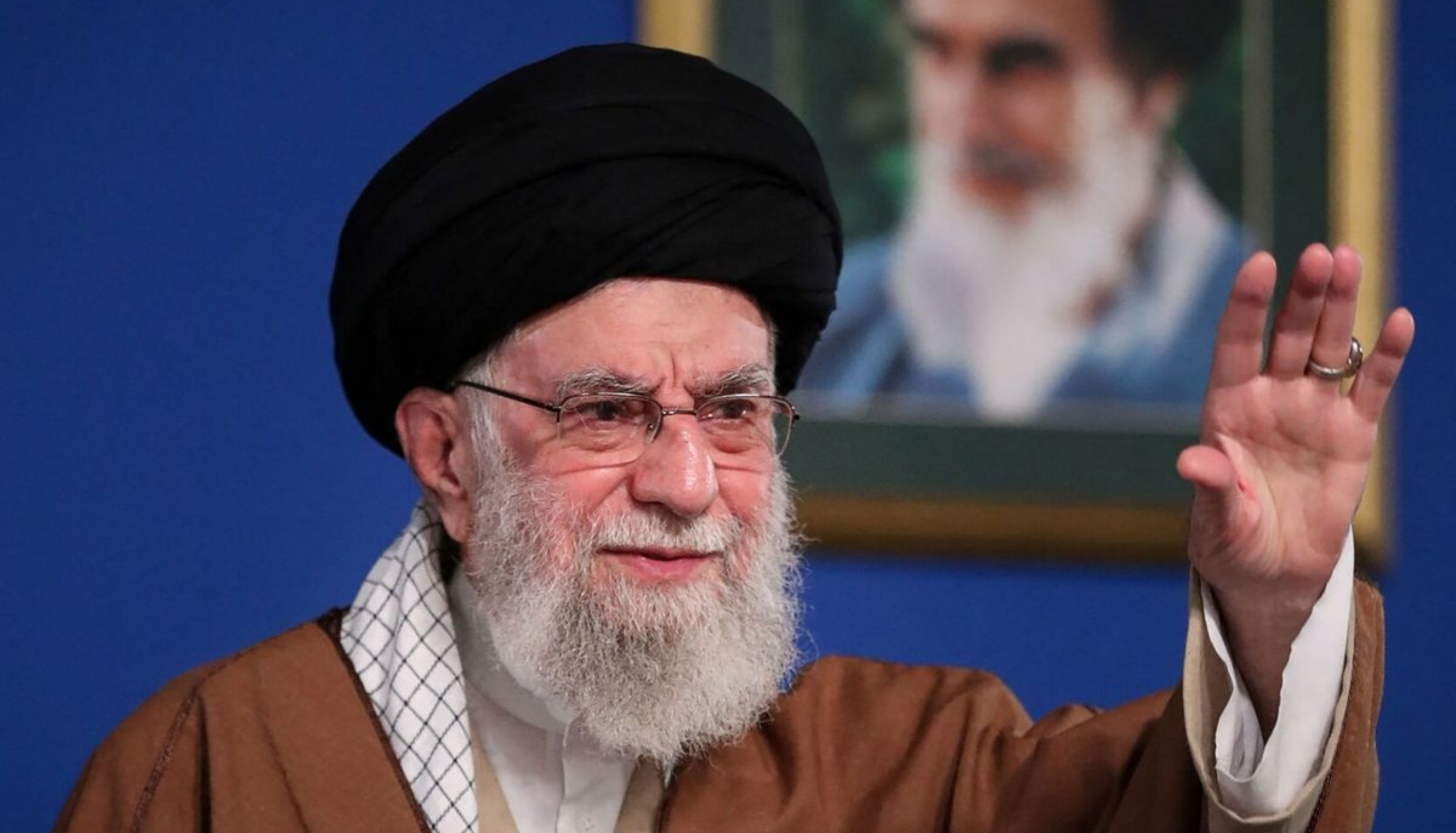 Elderly bearded man in a black turban and glasses raises his hand while speaking